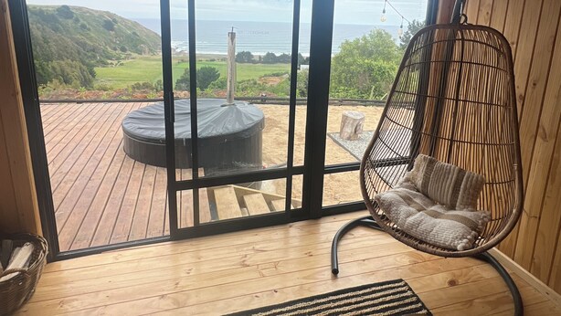 Un interior de madera con un amplio ventanal corredera que da a una terraza exterior. Una silla de mimbre con cojín está colocada cerca del ventanal.