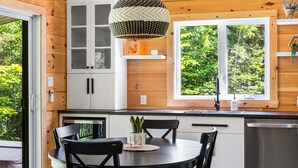 A modern kitchen with wooden cabinetry, a dining table with chairs, a pendant light, and a window with a view of greenery. - Spa, Nature & Riverside Escape | Ecureuil Roux (Trois-Rives)