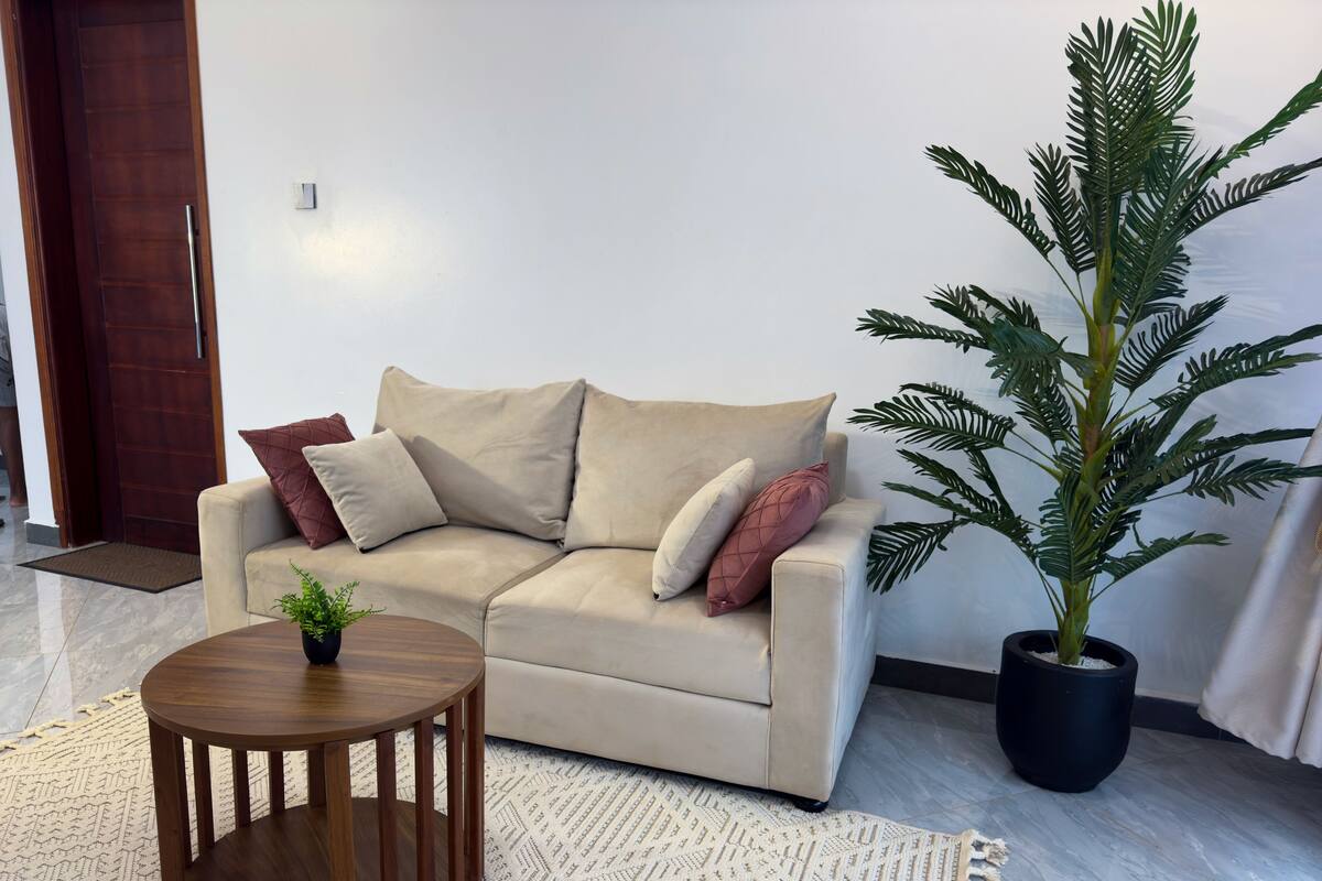 A beige sofa with multiple cushions, a small wooden side table, a potted plant, and a patterned rug in a living room.