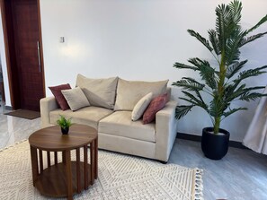A beige sofa with multiple cushions, a small wooden side table, a potted plant, and a patterned rug in a living room.