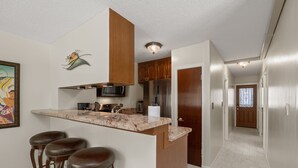 A kitchen with a marble countertop, stainless steel appliances, and wooden cabinets. There are three stools at the bar area. - Corner Unit 1 Block to Beach in Heart of Waikiki! FREE Parking & Wi-Fi, AC! (Honolulu)