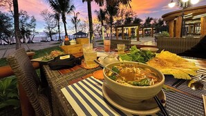 Un espace de restauration en plein air, avec une table dressée pour un repas, comprenant un bol de soupe et divers plats, sous un toit de chaume, offrant une vue sur le coucher du soleil et les palmiers.