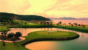 Un parcours de golf avec un obstacle d’eau, des palmiers et des montagnes qui se dessinent à l’horizon.