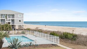 A two-story building with a swimming pool, a beach view, and a pier in the distance. - Oceanfront Condo w/ Pool, Late Summer Availability (Ocean Isle Beach)