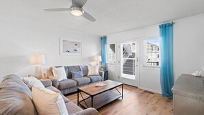 A living room with a sofa, coffee table, and two lamps. There is a door with a view of buildings outside. - Oceanfront Condo w/ Pool, Late Summer Availability (Ocean Isle Beach)