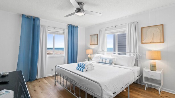 A bedroom with a bed, two bedside tables, a ceiling fan, and a view of the beach. - Oceanfront Condo w/ Pool, Late Summer Availability (Ocean Isle Beach)