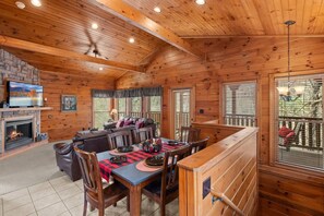 Interior acolhedor de uma cabana com lareira, televisão, área de jantar e uma grande janela com vista para um deck.