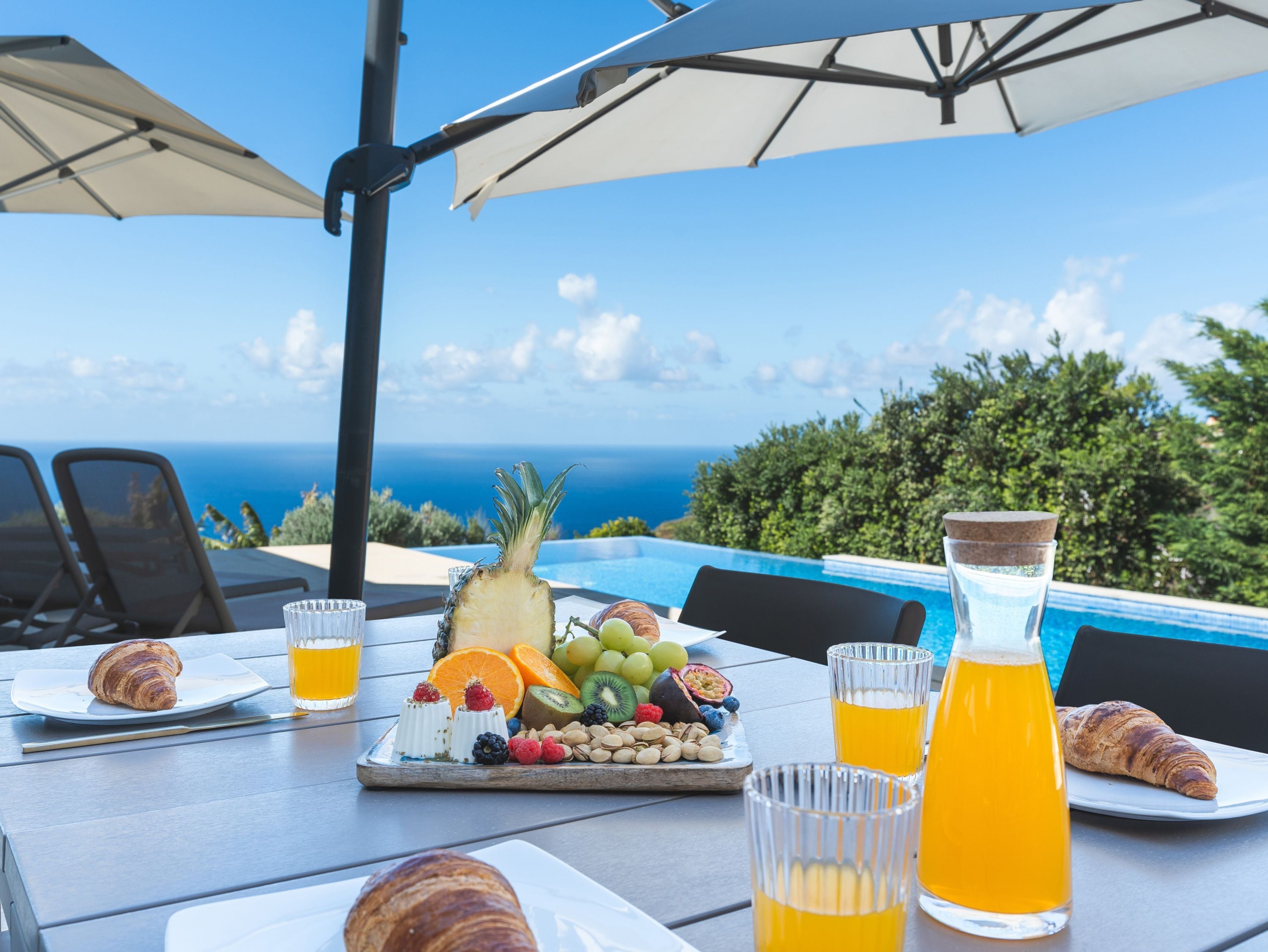 Desayuno con cruasanes, frutas, frutos secos y zumo de naranja sobre una mesa con vistas a la piscina.