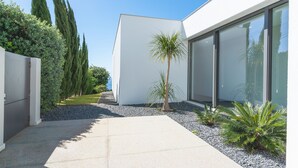 Um edifício moderno com fachada clara e branca, grandes janelas e um jardim bem cuidado, com diversas plantas e um caminho de brita.