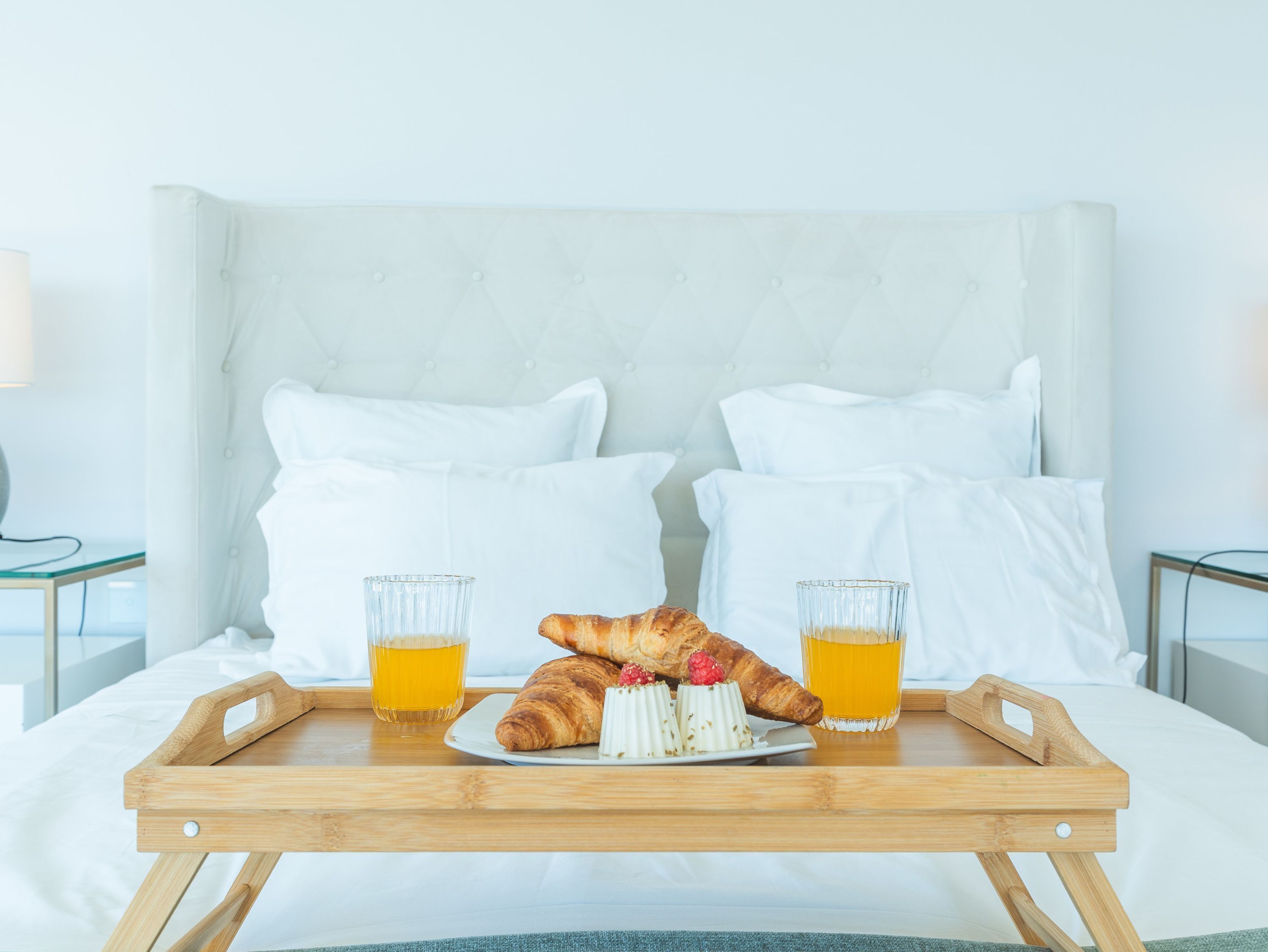 A bed with a wooden tray holding a croissant, two glasses of orange juice, and a plate with a pastry and raspberries.