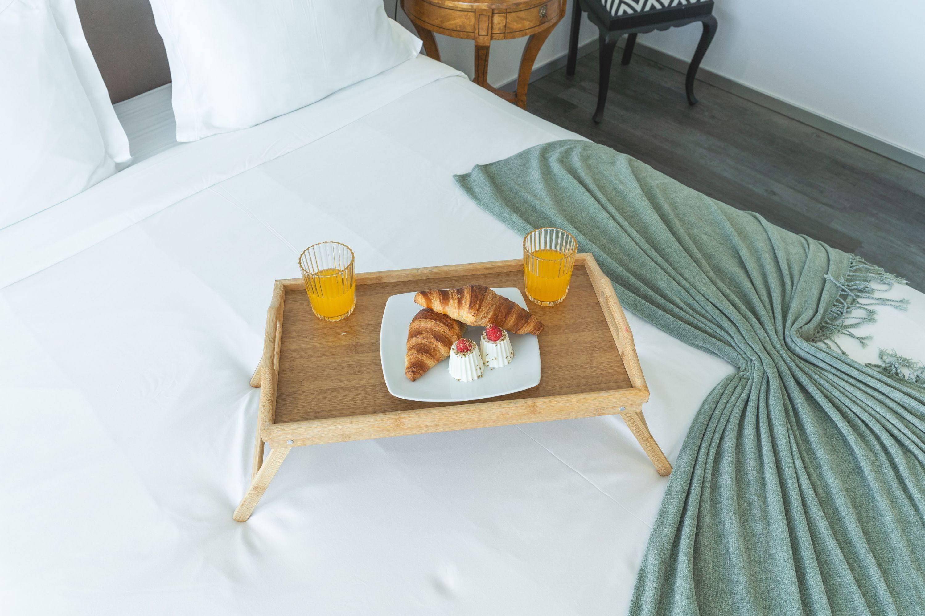 A neatly made bed with white linens, a wooden tray with croissants and orange juice, and a green throw blanket.