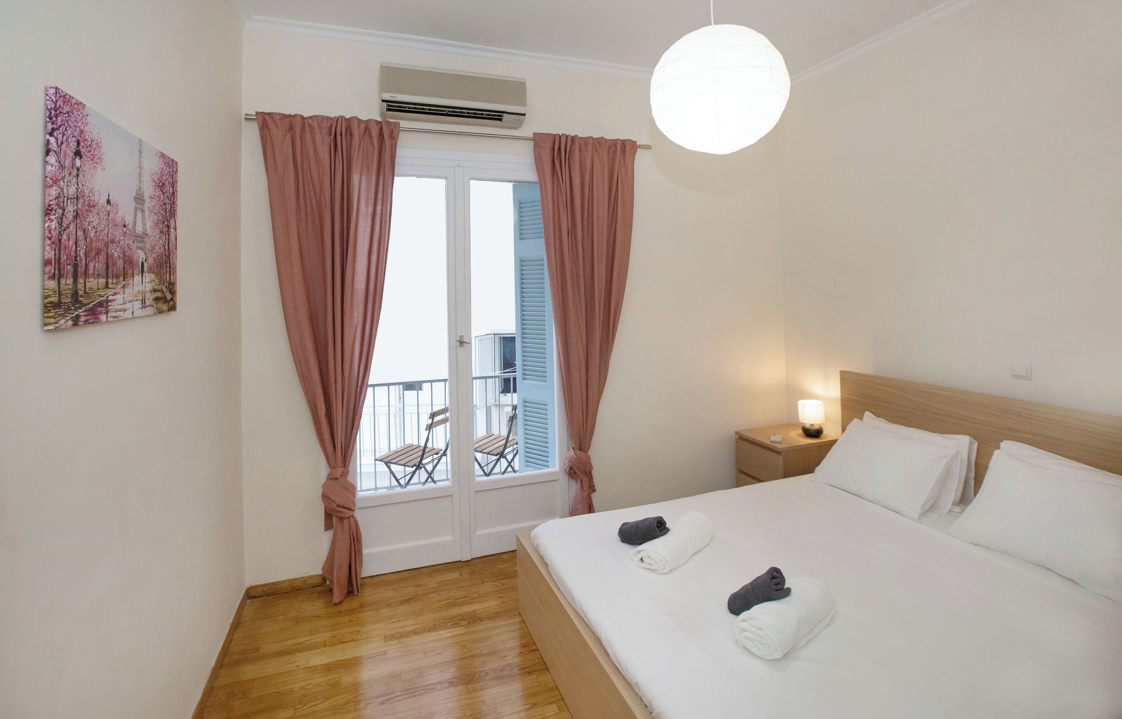 Ein Hotelzimmer mit Bett, Nachttisch, Nachttischlampe, einem Fenster mit Vorhängen, einer Klimaanlage, einem Balkon mit Stühlen und einem Bild an der Wand.