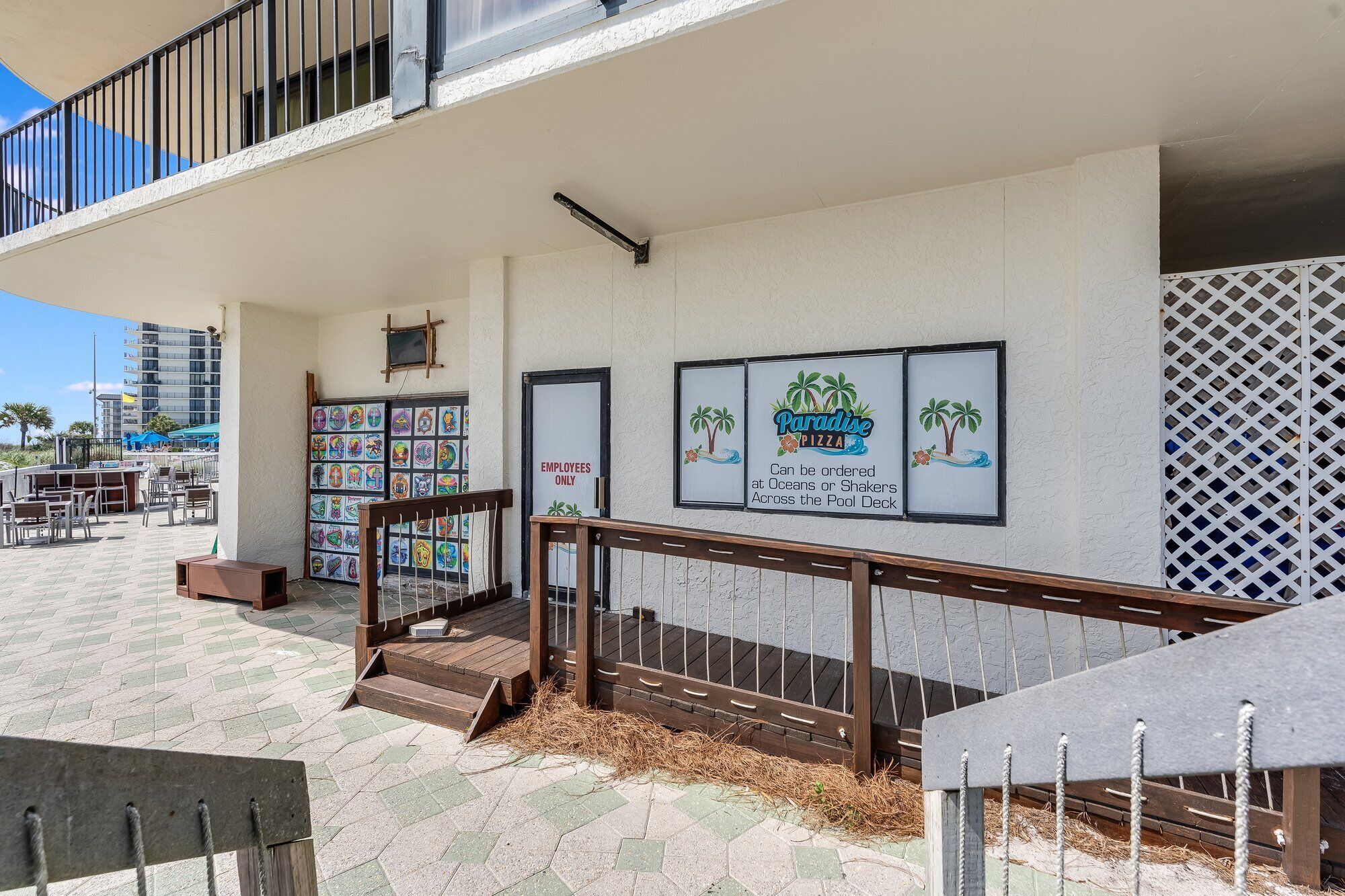 Un bâtiment situé en bord de mer, doté d’une terrasse avec piscine, de sièges en plein air et d’une enseigne publicitaire pour la pizzeria « Paradise Pizza ».
