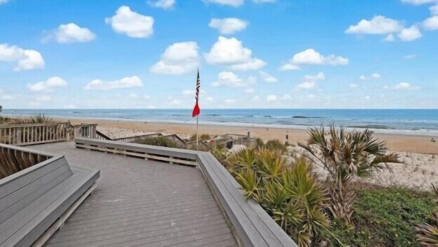 Une promenade en bois mène à une plage où se trouve un mât de drapeau au centre.