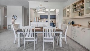 Un coin repas avec une table en bois et des chaises blanches, un luminaire suspendu et une cuisine en arrière-plan.