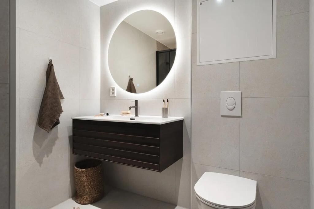 Un baño moderno con inodoro blanco, tocador oscuro con espejo y un espejo redondo con luz.