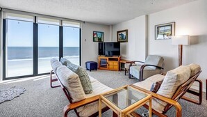 A spacious living room with a large window overlooking the ocean, a flat-screen TV, and a comfortable seating area with a coffee table. - Crescent Breeze - Oceanfront w/incredible views, lanai & pool (St. Augustine)