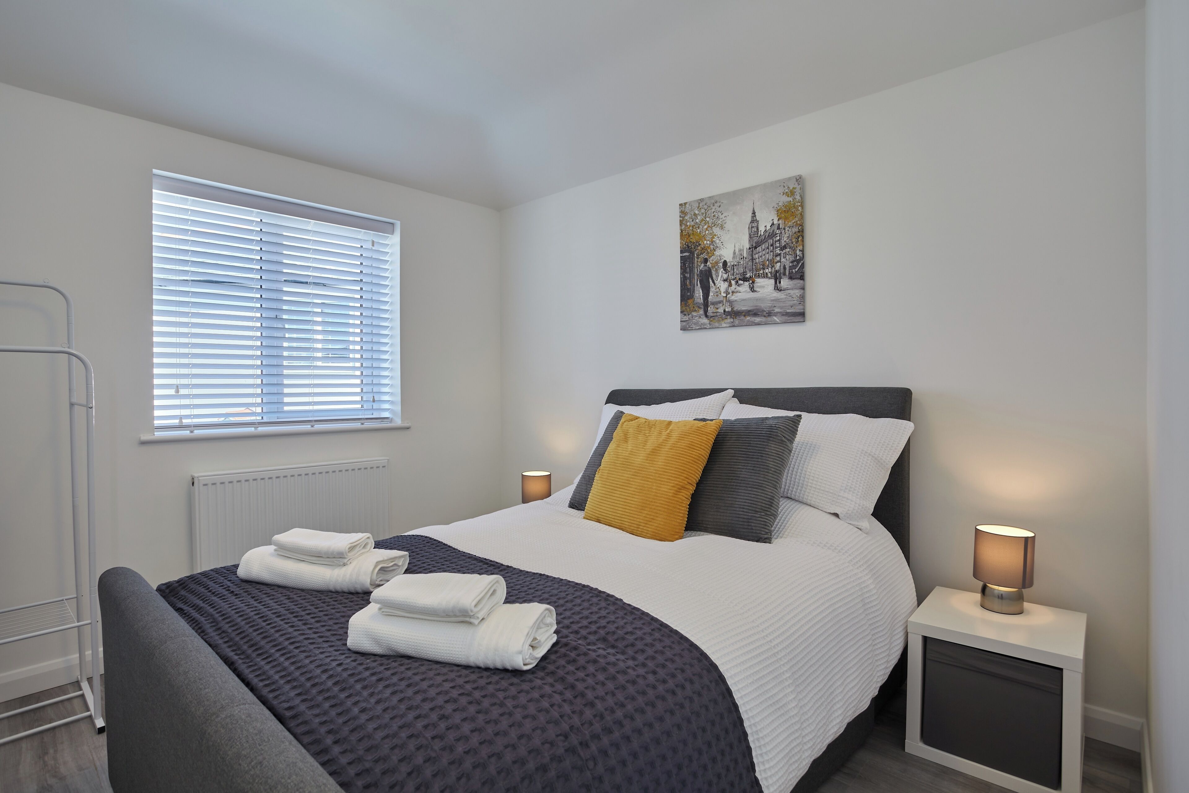 A modern bedroom with a bed, bedside lamps, a radiator, and a window with blinds.
