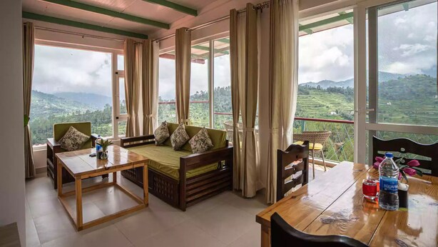 Una habitación con una mesa de madera, un sofá con cojines y grandes ventanas que dan a un paisaje montañoso.