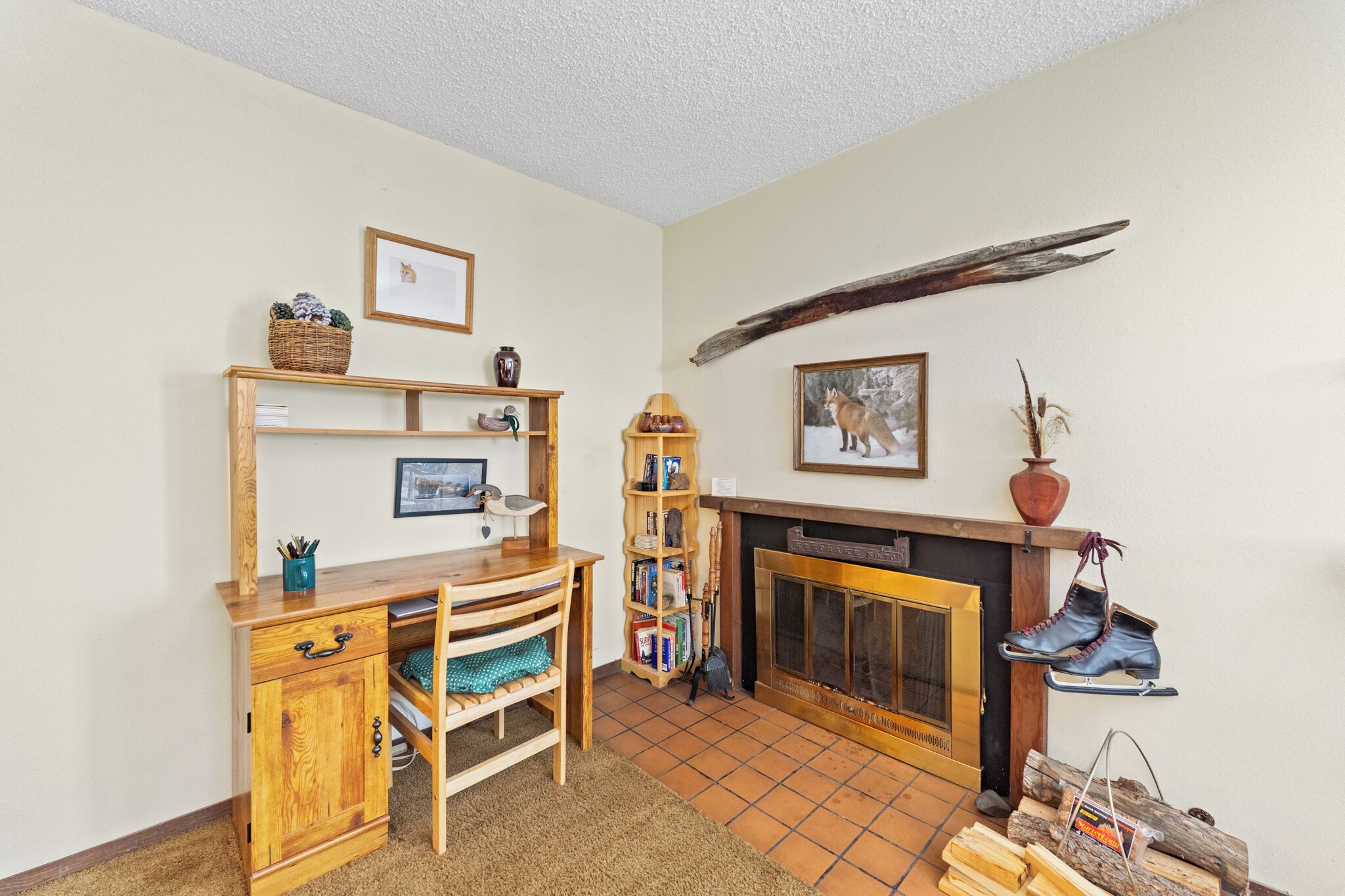 A cozy living room with a wooden desk, bookshelves, a fireplace, and a pair of boots hanging on the wall.