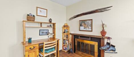 A cozy living room with a wooden desk, bookshelves, a fireplace, and a pair of boots hanging on the wall.