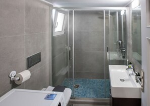 A modern bathroom with a glass-enclosed shower, a white sink, and a toilet with a paper towel dispenser. - Modern Rental with Cozy Charm by GHH (Athina)