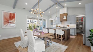 Una cocina moderna con armarios blancos, una isla de madera, iluminación colgante y un comedor con sillas blancas y una mesa preparada para seis personas.