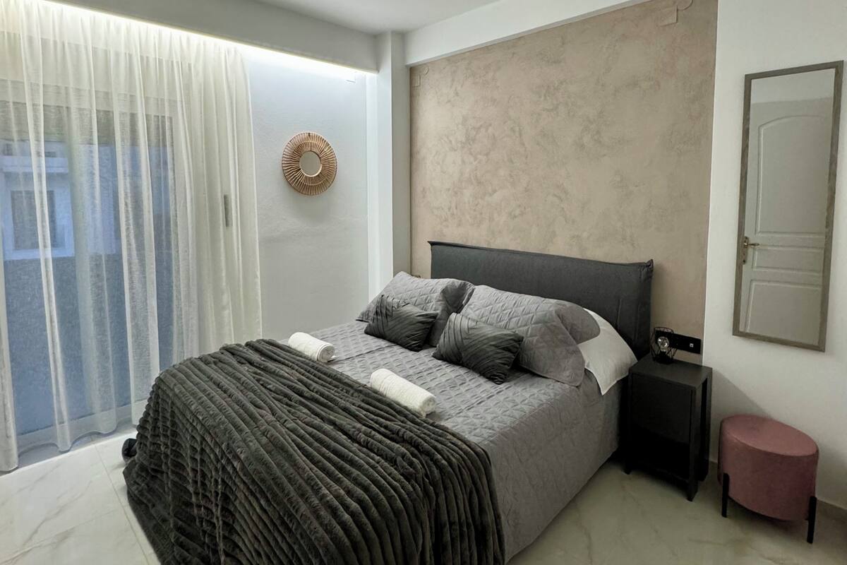 Un dormitorio moderno con una cama grande, una mesita de noche, un taburete pequeño y un espejo fijado a la pared.