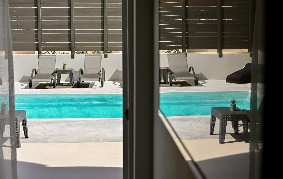 A poolside area with lounge chairs and tables, a view of the ocean, and a building with horizontal slats.