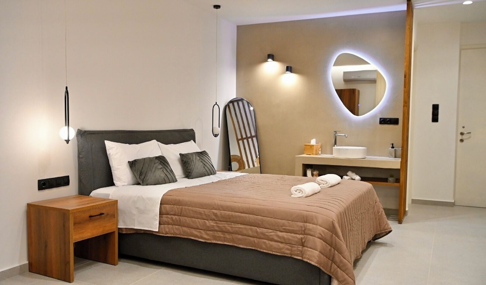 Un dormitorio moderno con cama, mesita de noche y baño con lavamanos y espejo.