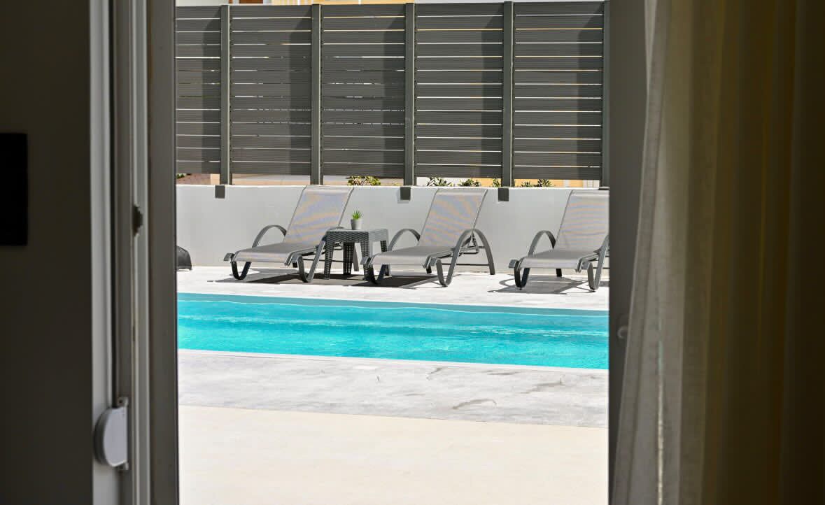 A poolside area with lounge chairs and a table, viewed through an open door.