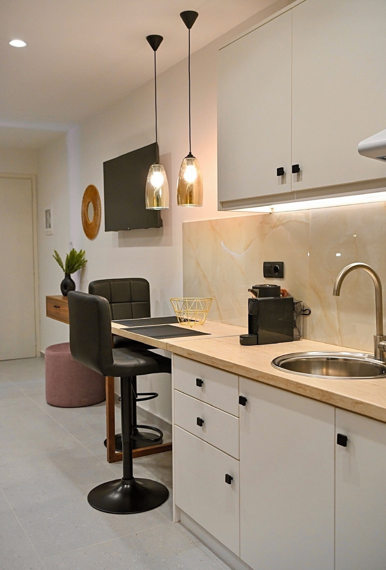 A modern kitchen with a marble countertop, stainless steel sink, and black cabinets. There are pendant lights, a coffee machine, and a plant on the counter.