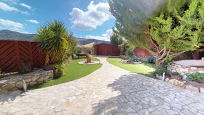 Ein gepflegter Garten mit unterschiedlichen Pflanzen umgibt ein rotes Gebäude, auf das ein Steinweg führt, begrenzt durch einen Holzzaun.