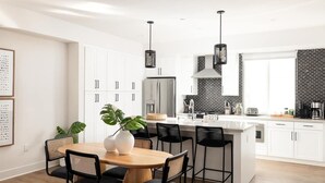 Une cuisine moderne avec des meubles blancs, un îlot central, des chaises noires et des luminaires suspendus.