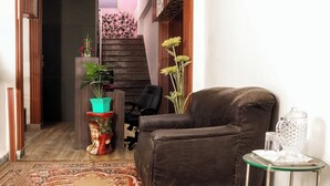 A living room with a brown leather sofa, a small glass table with a crystal pitcher, and a colorful vase with flowers. A staircase with a pink railing and a wooden wall is visible in the background. - Hotel Sunrise Inn (Noida)