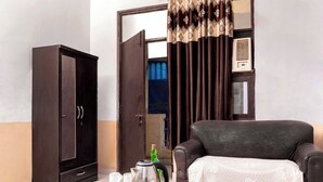 A room with a dark brown wardrobe, a sofa with a white blanket, a glass table with a kettle and cups, and an air conditioning unit on the wall. - Hotel Sunrise Inn (Noida)