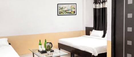A hotel room with two beds, a glass table with bottles and a kettle, a framed picture on the wall, and a door with a patterned curtain.