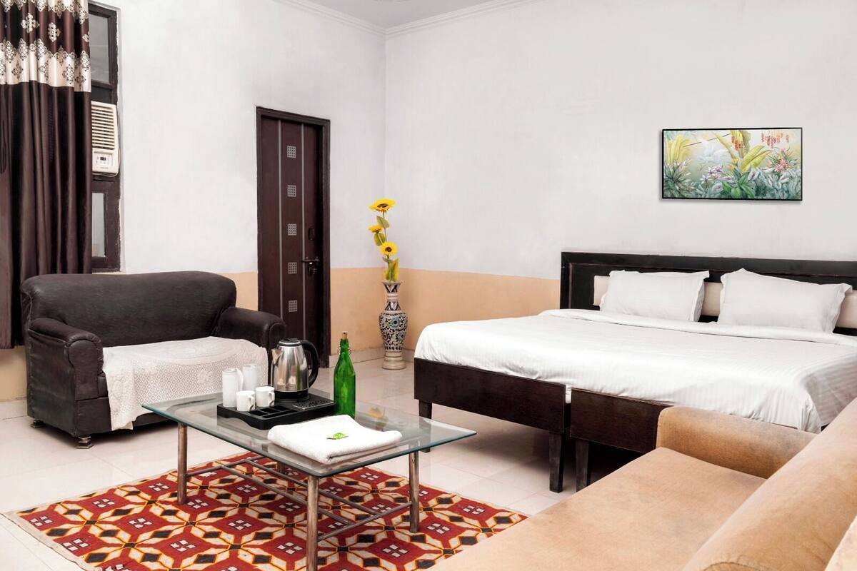 Habitación de hotel con una cama, un sofá, una mesa de centro de vidrio, un jarrón con flores y un cuadro en la pared.