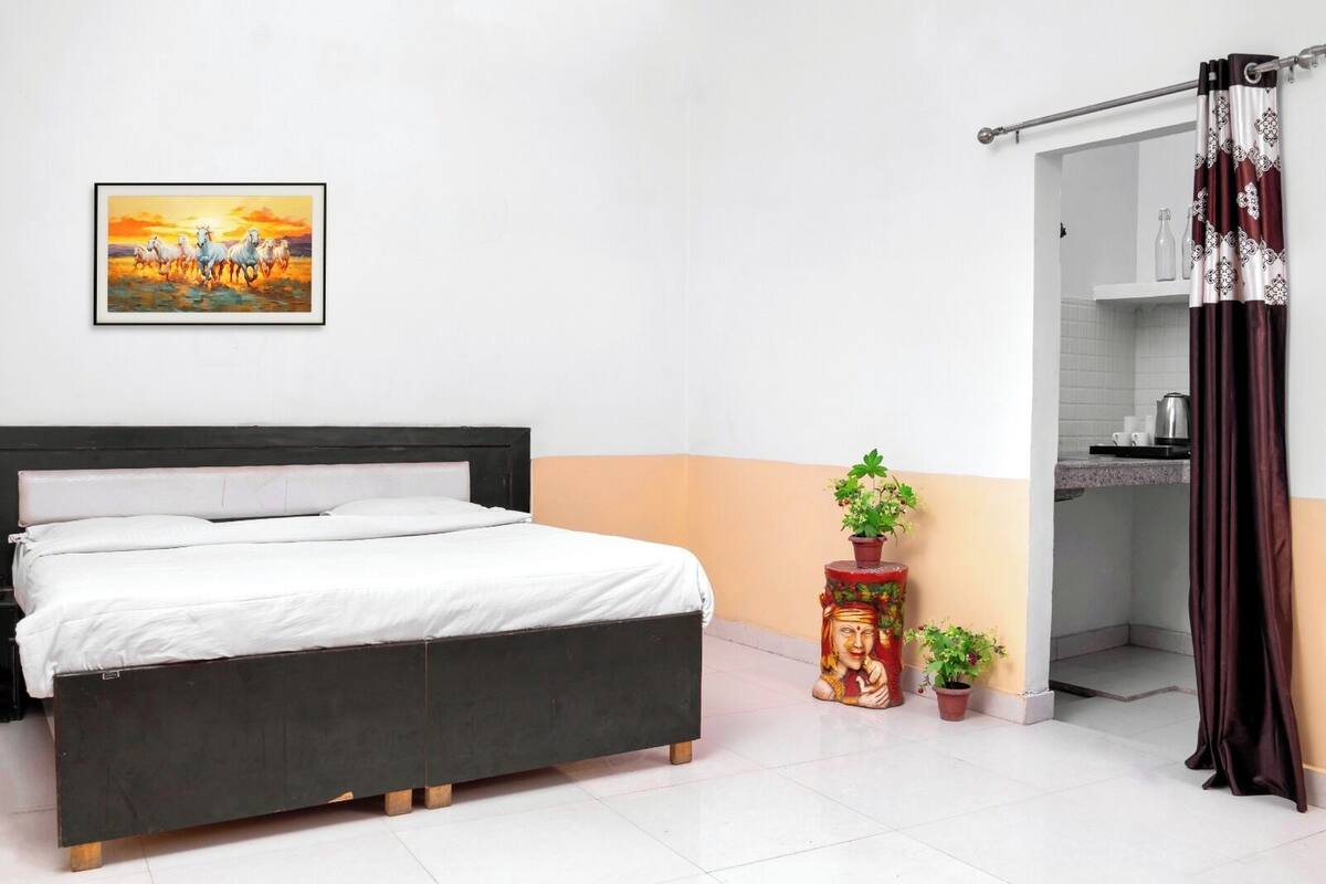 Un dormitorio con cama, un cuadro, una mesita con plantas y un baño visible a través de una puerta abierta.