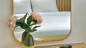 Un dormitorio con un espejo grande, una cama con ropa de cama blanca y un florero con flores sobre una mesita de noche.