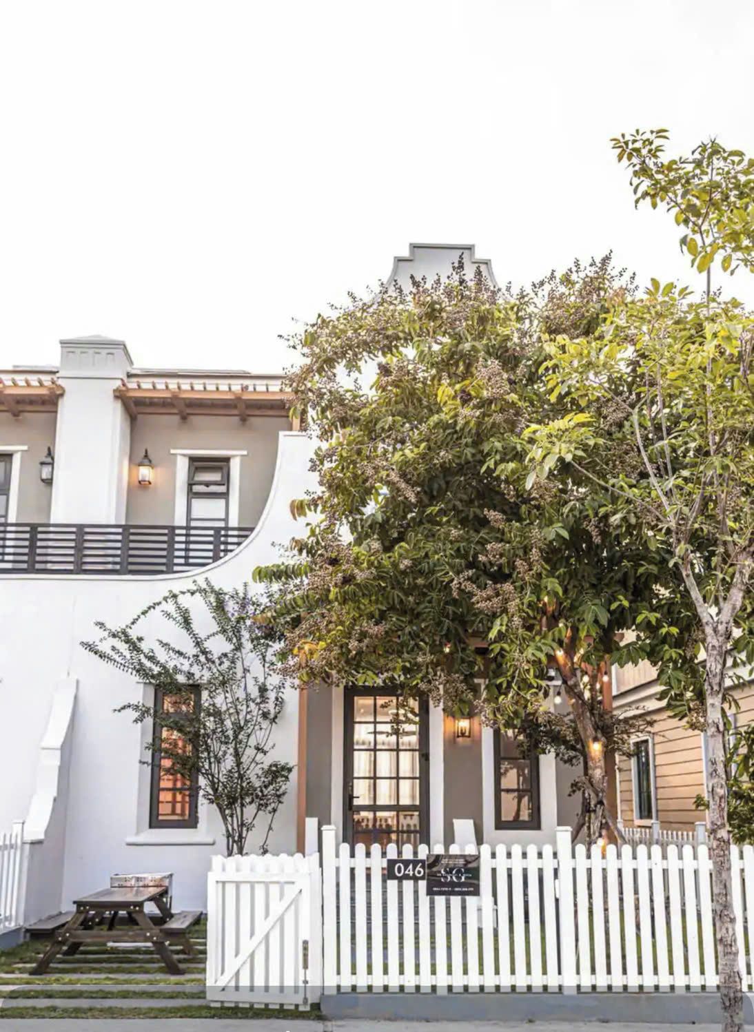 Un edificio de dos pisos con fachada blanca, una cerca de piquetes blancos y un árbol en primer plano.