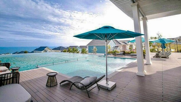 Zona de piscina con sillones de descanso y sombrillas, con vistas a un cuerpo de agua y edificios.