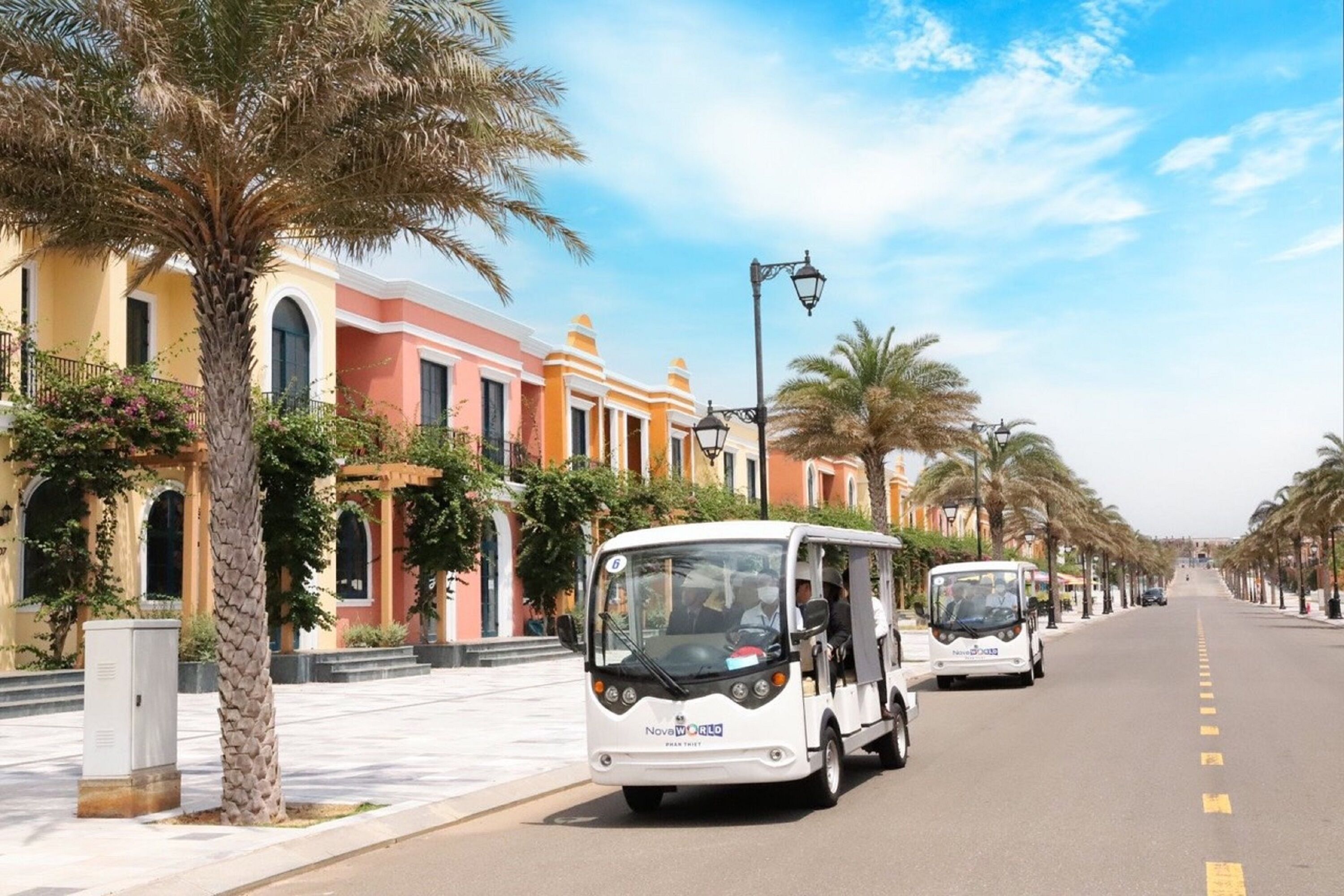 A street lined with palm trees and colorful buildings, with a white bus in the foreground.