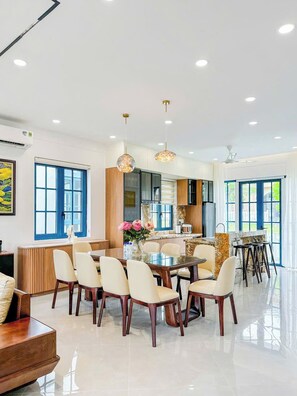 A spacious dining area with a round table and chairs, a kitchen in the background, and large windows. - Novaworld Phan Thiet - QT Villa (Tien Thanh)