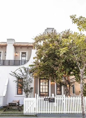 A two-story building with a white facade, a white picket fence, and a tree in the foreground. - Novaworld Phan Thiet - QT Villa (Tien Thanh)