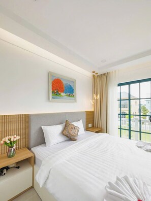 A hotel room with a bed, a nightstand, a vase of flowers, a painting, and a window with a view. - Novaworld Phan Thiet - QT Villa (Tien Thanh)