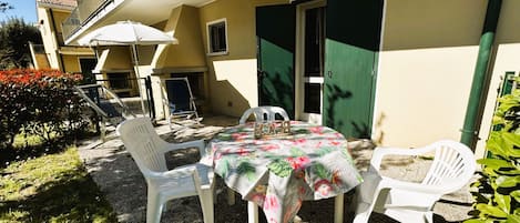 An outdoor patio with a table and chairs, a white umbrella, and a door with green shutters.