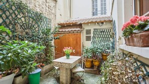 A courtyard with a wooden table and chairs, surrounded by potted plants and a stone wall. - Superbe Apartment Le Marais/Place des Vosges (Paris)