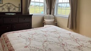 A bedroom with a bed, a dresser, a chair, and two windows with curtains. - Darnley Dream vacation home perfect to experience PEI to its fullest. (Kensington)