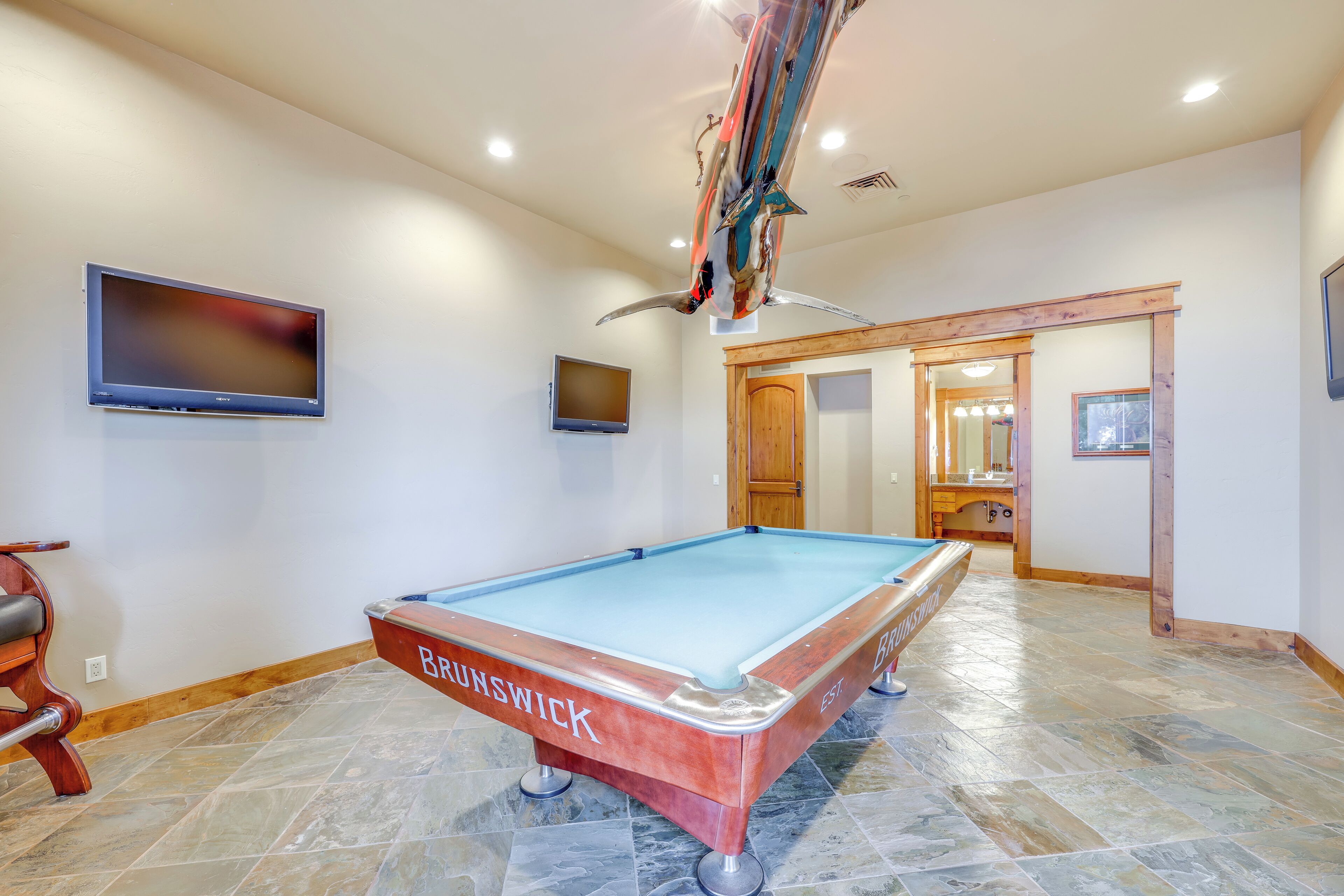 A pool table labeled 'BRUNSWICK' in a room with tiled flooring, a mounted television, and a wooden door leading to another area.